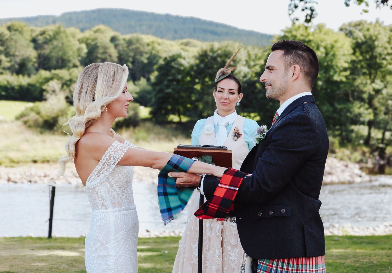 A couple exchanges vows during an outdoor wedding ceremony near a river with mountains in the background.