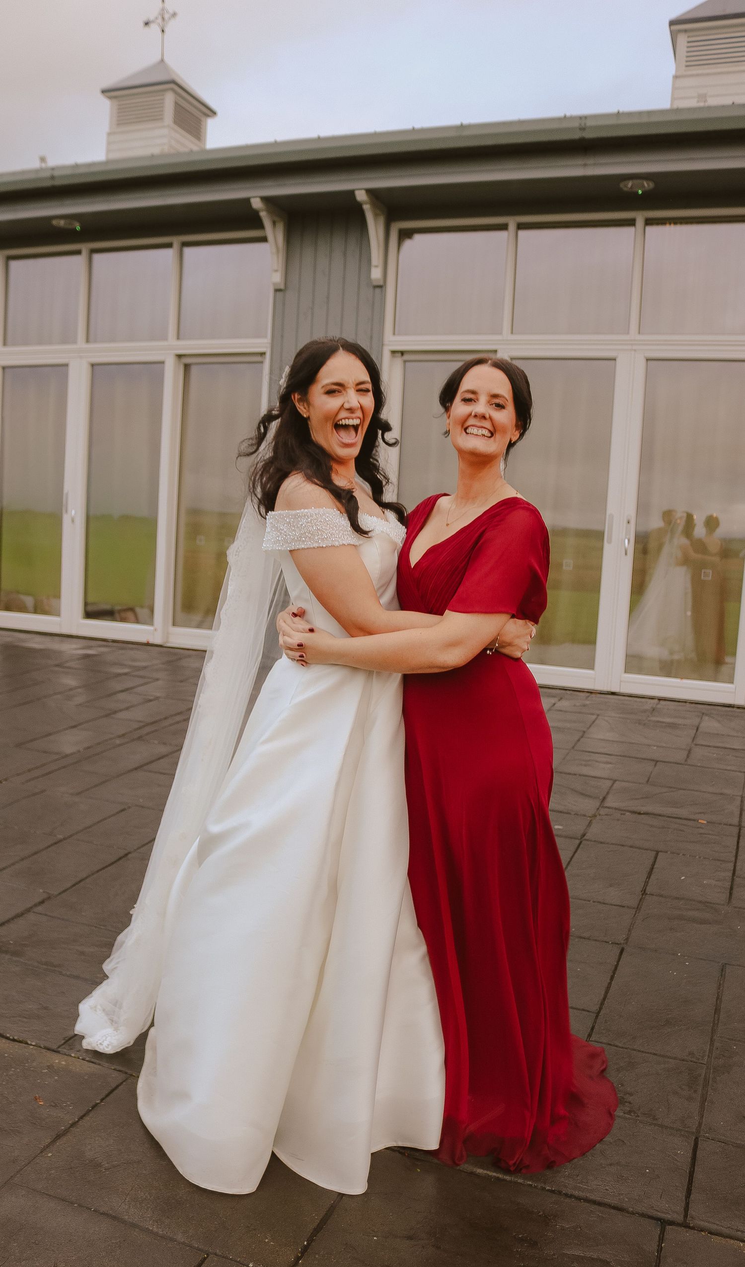 Two women in contrasting white and red dresses share joyful laughter on a stone patio during a special occasion.
