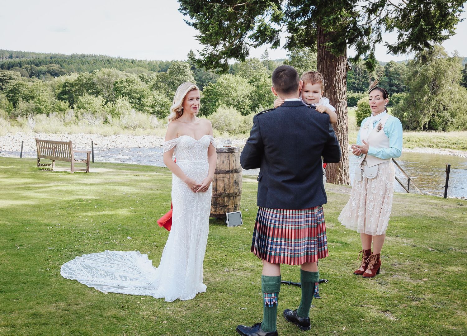 Wedding ceremony participants gather on a grassy lawn overlooking a scenic waterfront landscape.