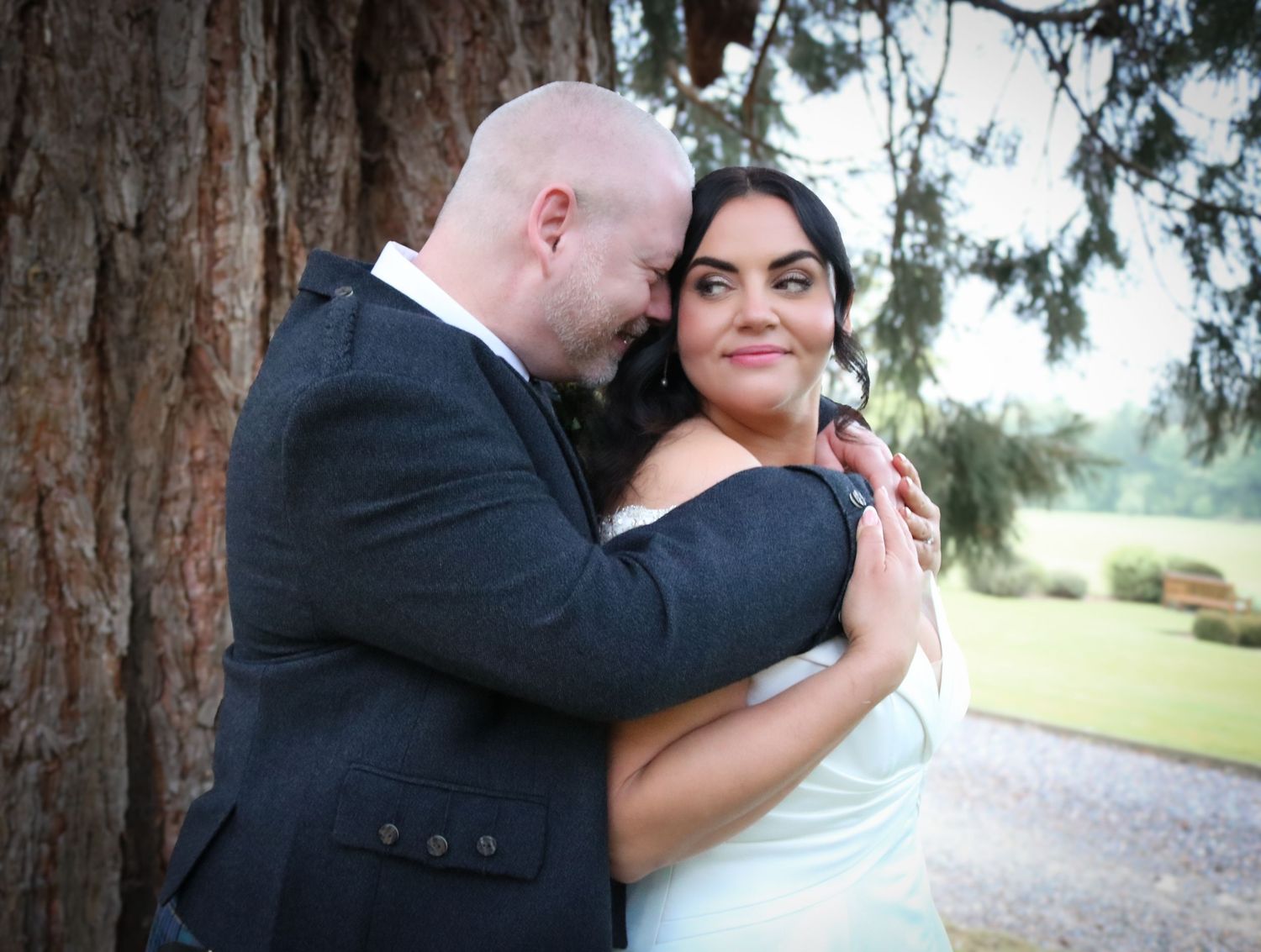 A bride and groom share a romantic embrace near a large tree outdoors on their wedding day.
