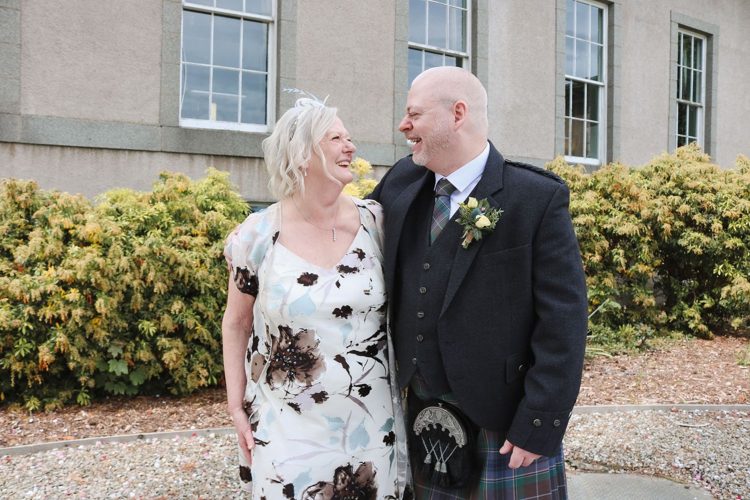 A smiling couple in wedding attire, woman in floral dress and man in Scottish kilt, standing outside a stone building.