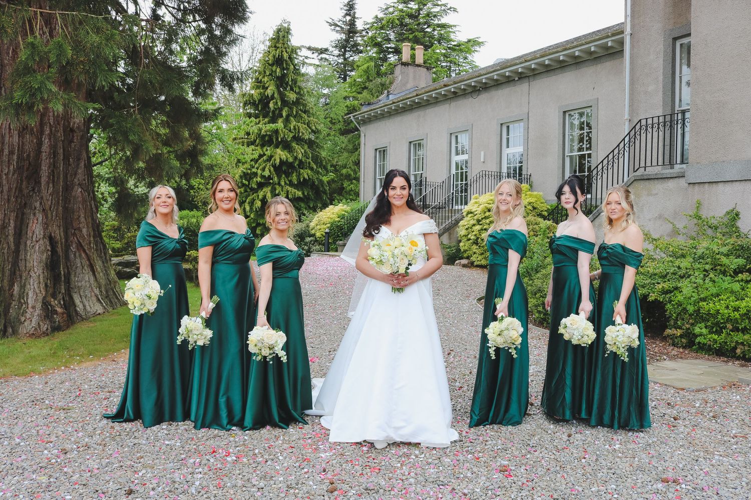 Bride in white gown with six bridesmaids in emerald green off-shoulder dresses holding white bouquets outside elegant estate.
