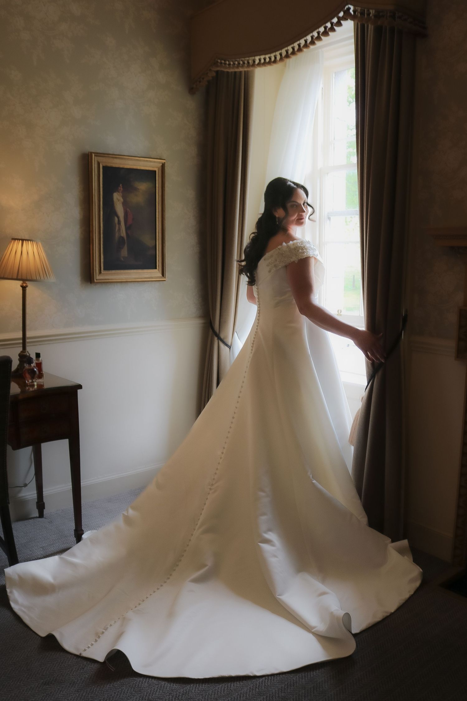 Bride in elegant white gown with long train stands by window in ornate room, soft natural light illuminating her silhouette.