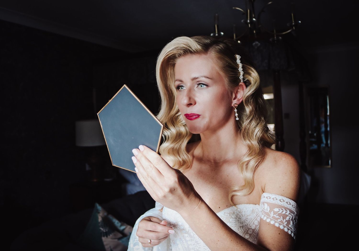A woman in a white off-shoulder dress with blonde curls holds a small mirror in a dimly lit room.