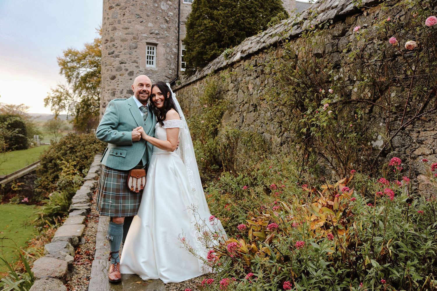 A romantic couple shares intimate moments among stone buildings and lush gardens in a rustic countryside setting.