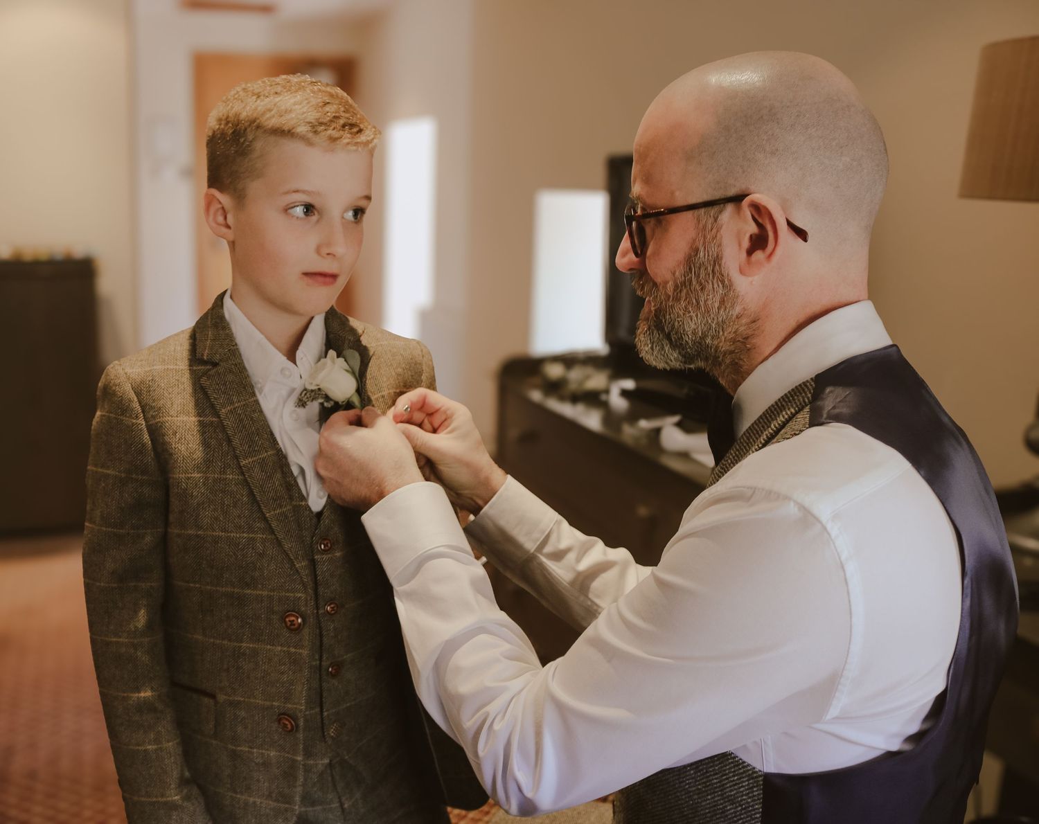 A well-dressed man in a vest and glasses adjusts a young boy's tie in a formal setting.