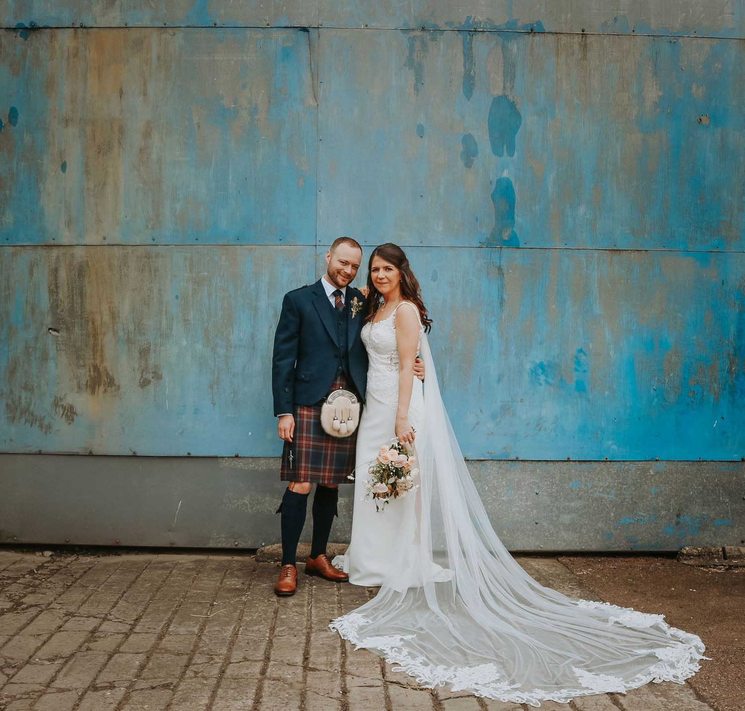 wedding-photographer-aberdeen- Newlywed couple poses against industrial blue wall in artistic wedding portraits with flowing white veil.
