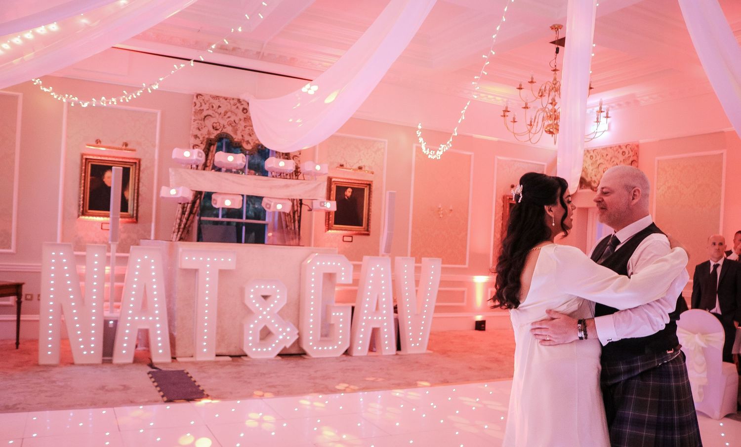 Bride and groom share first dance in front of illuminated NAT&GAV marquee letters at elegant pink wedding reception.