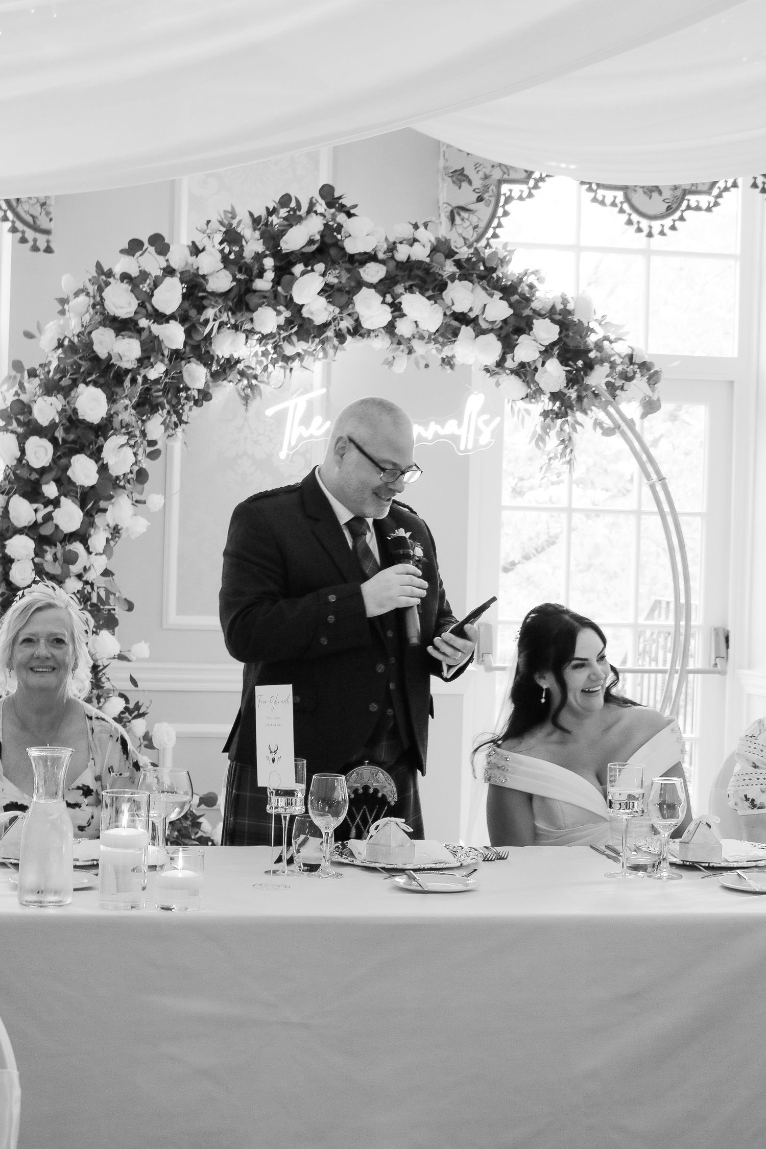 Black and white wedding reception photo of groom giving speech at head table with floral arch backdrop.