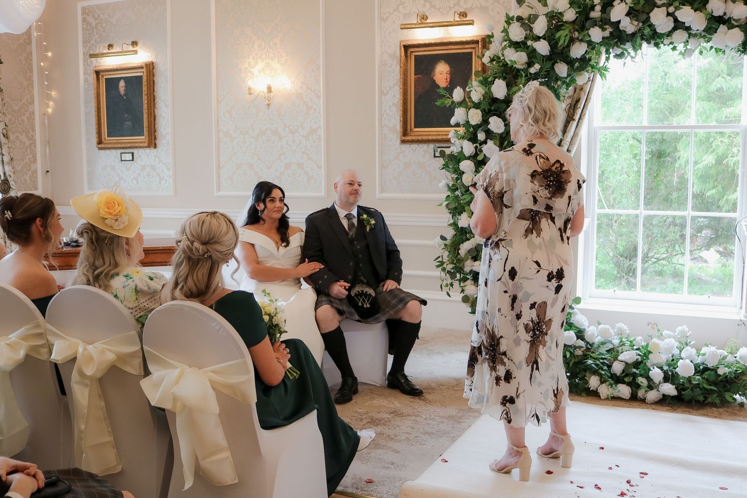 Elegant wedding ceremony in a white room with floral arch, bride in white gown, groom in kilt, and guests seated on chairs.