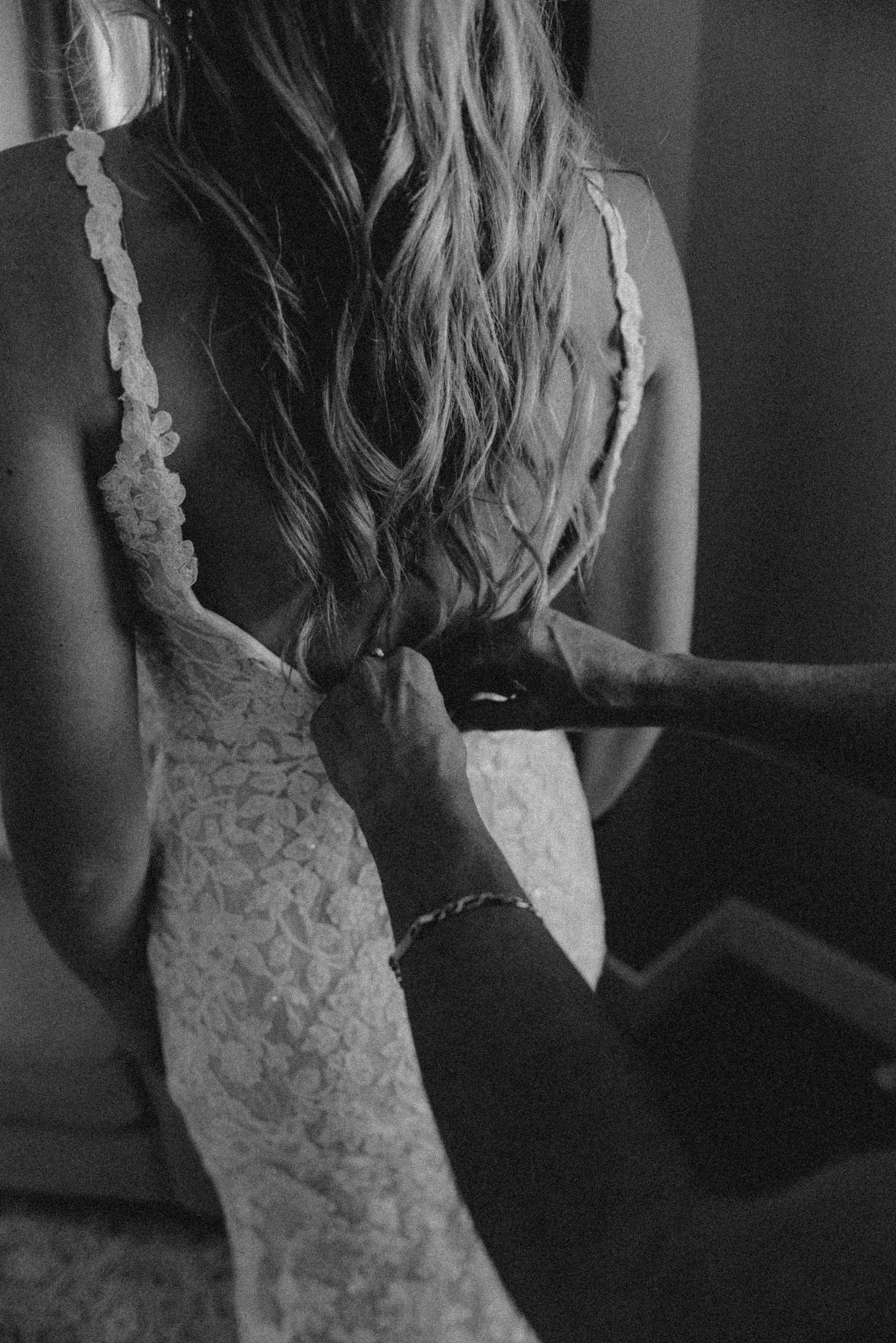 Black and white photo of someone helping zip up a delicate lace wedding dress with long flowing hair.