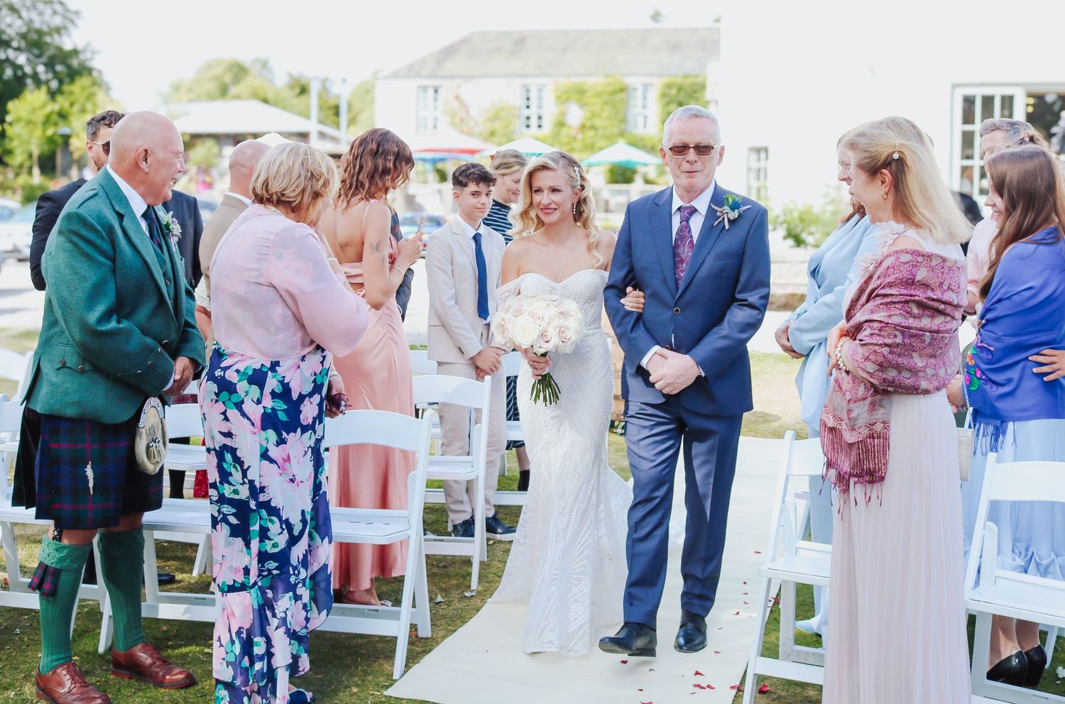 A group of wedding guests stand together during an outdoor ceremony with white chairs and green grass.
