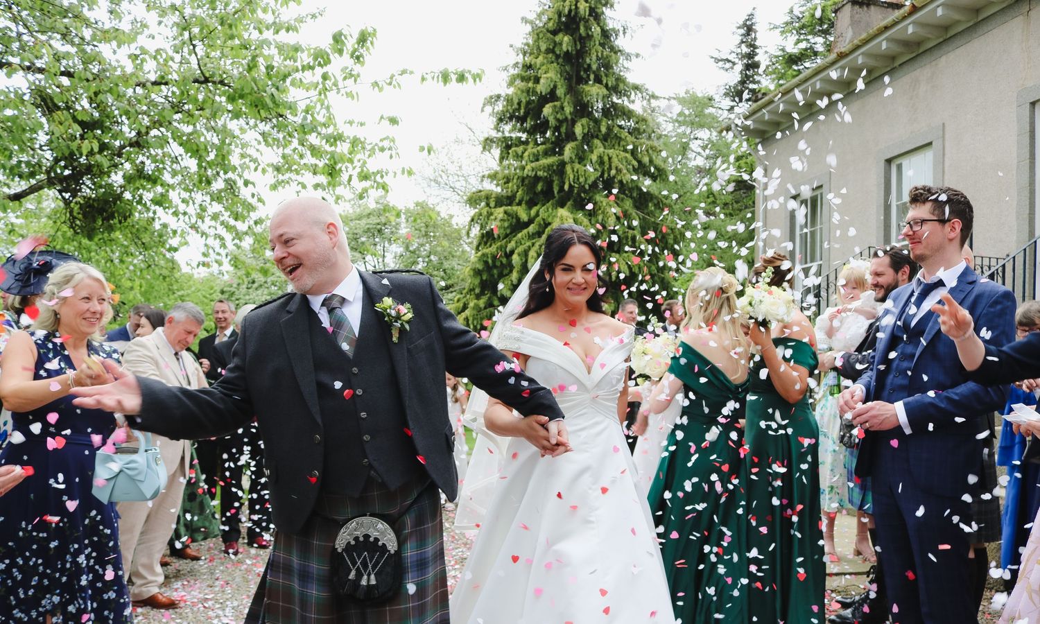 Bride and groom walk through confetti shower with guests celebrating at outdoor Scottish wedding ceremony.