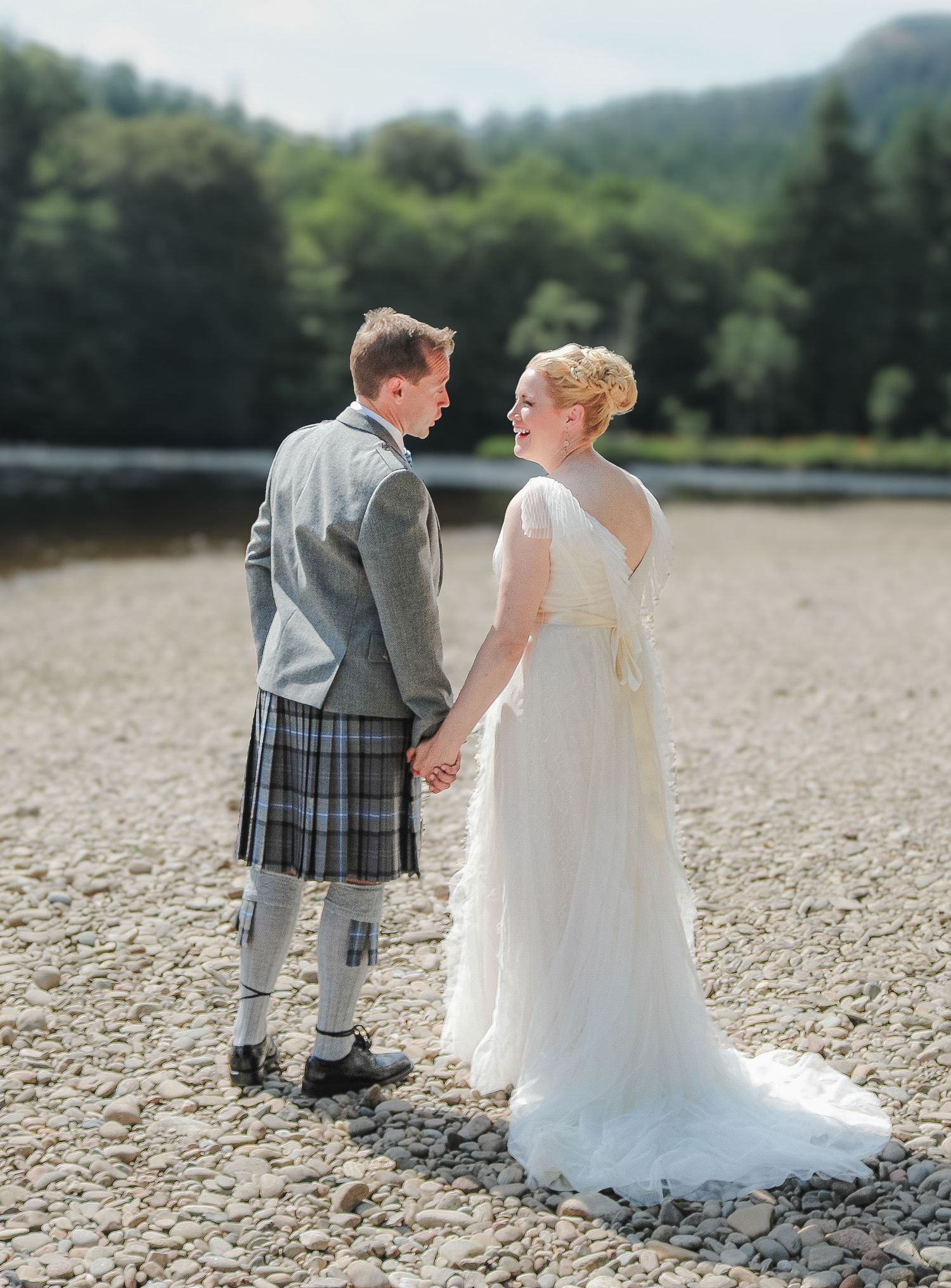 A couple in wedding attire stands on a rocky beach, with the groom wearing a traditional Scottish kilt and grey jacket.