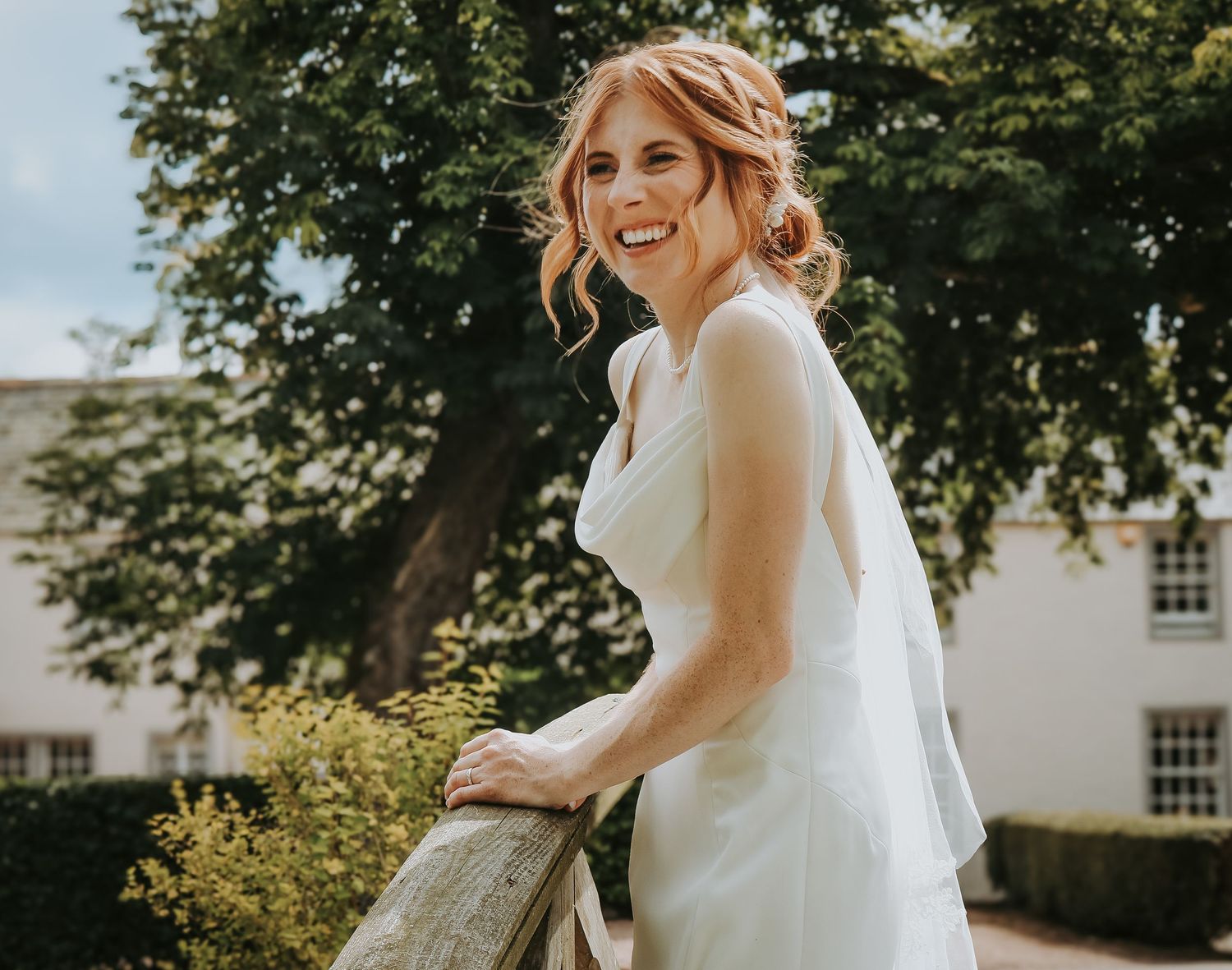 A bride in a flowing white wedding dress poses outdoors in a garden setting with natural sunlight.