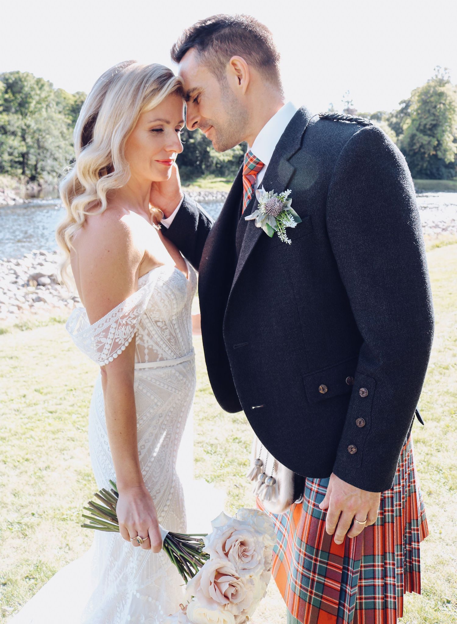 Romantic wedding couple in traditional Scottish attire with groom wearing kilt embrace outdoors on sunny day.