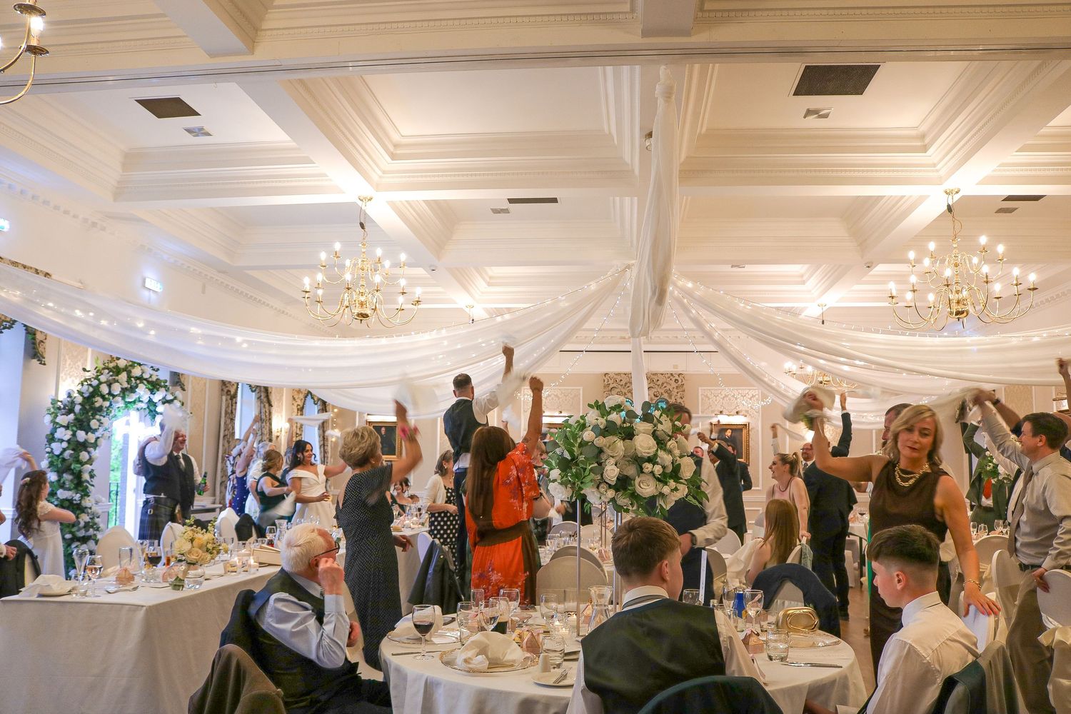 Elegant wedding reception in a grand ballroom with guests celebrating at round tables adorned with floral centerpieces.