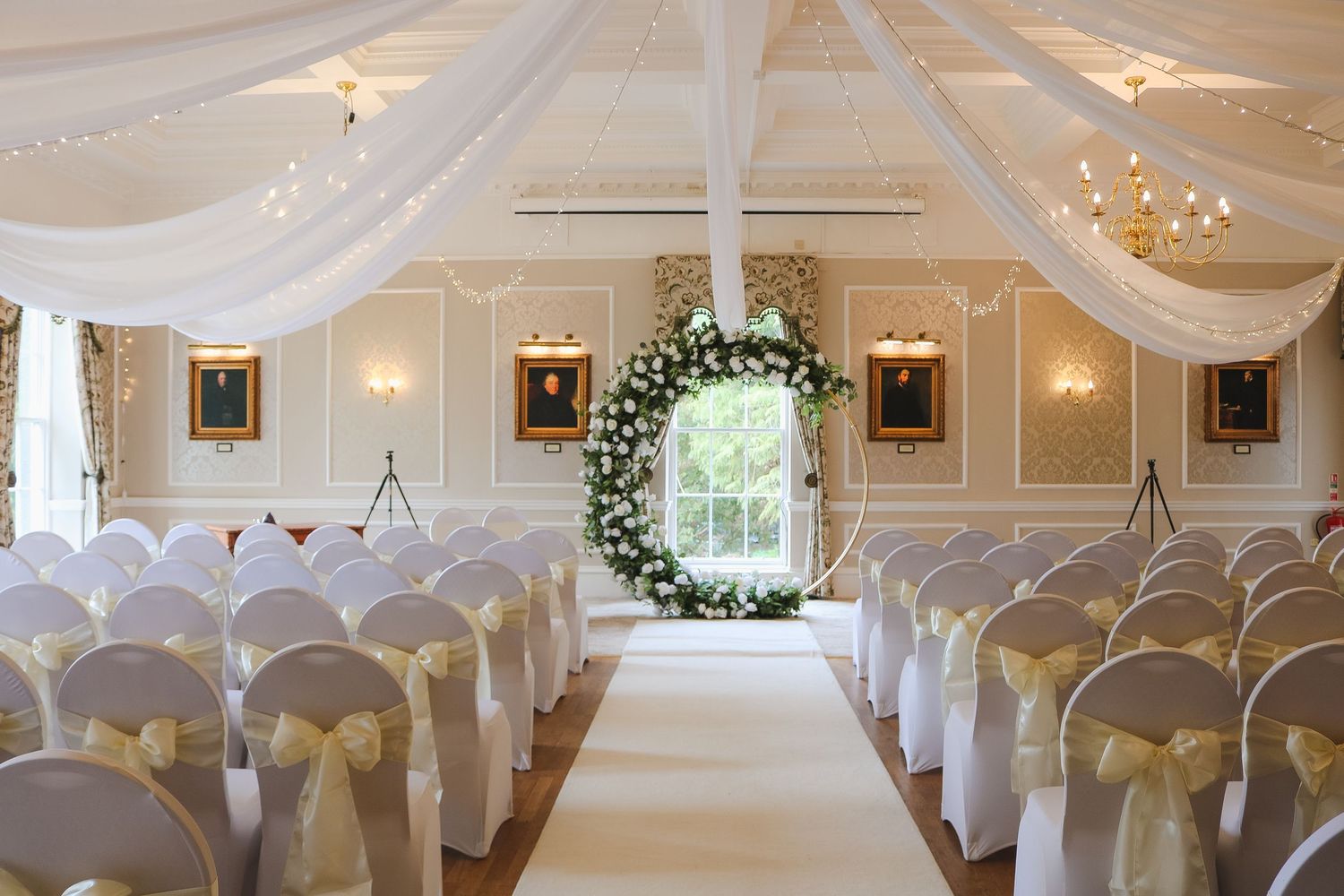 Elegant wedding ceremony venue with white draped ceiling, floral arch, and chairs adorned with gold bows lining the aisle.