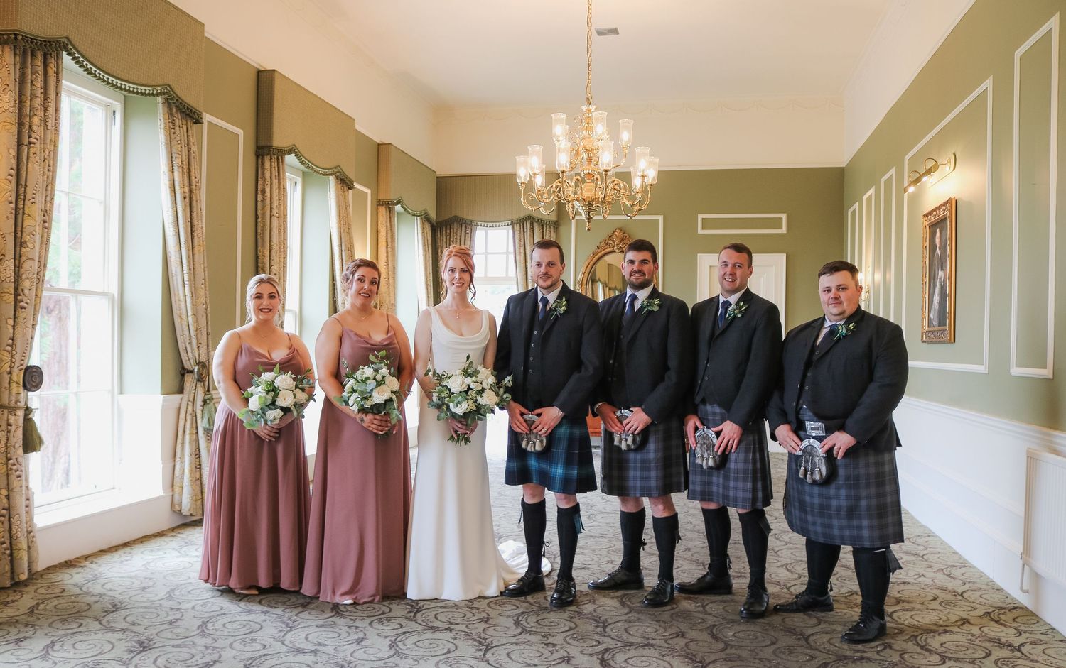 Wedding party wearing dusty rose bridesmaid dresses and traditional Scottish kilts poses in an elegant room with chandeliers.