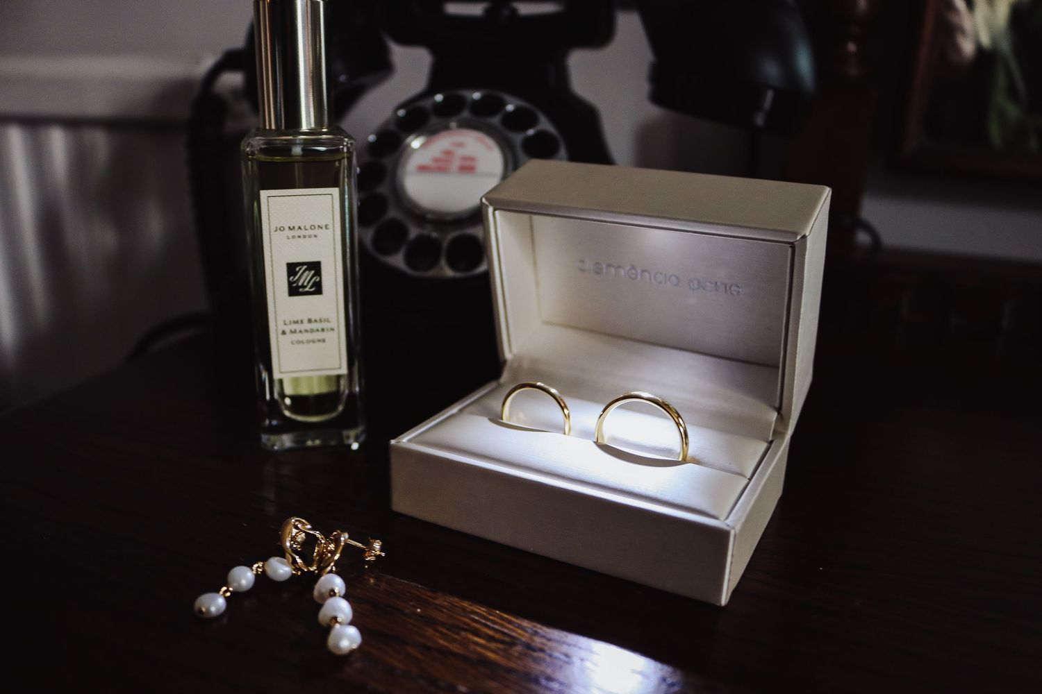 Elegant arrangement of Jo Malone perfume, glasses in a display case, and pearl drop earrings on dark wooden surface.