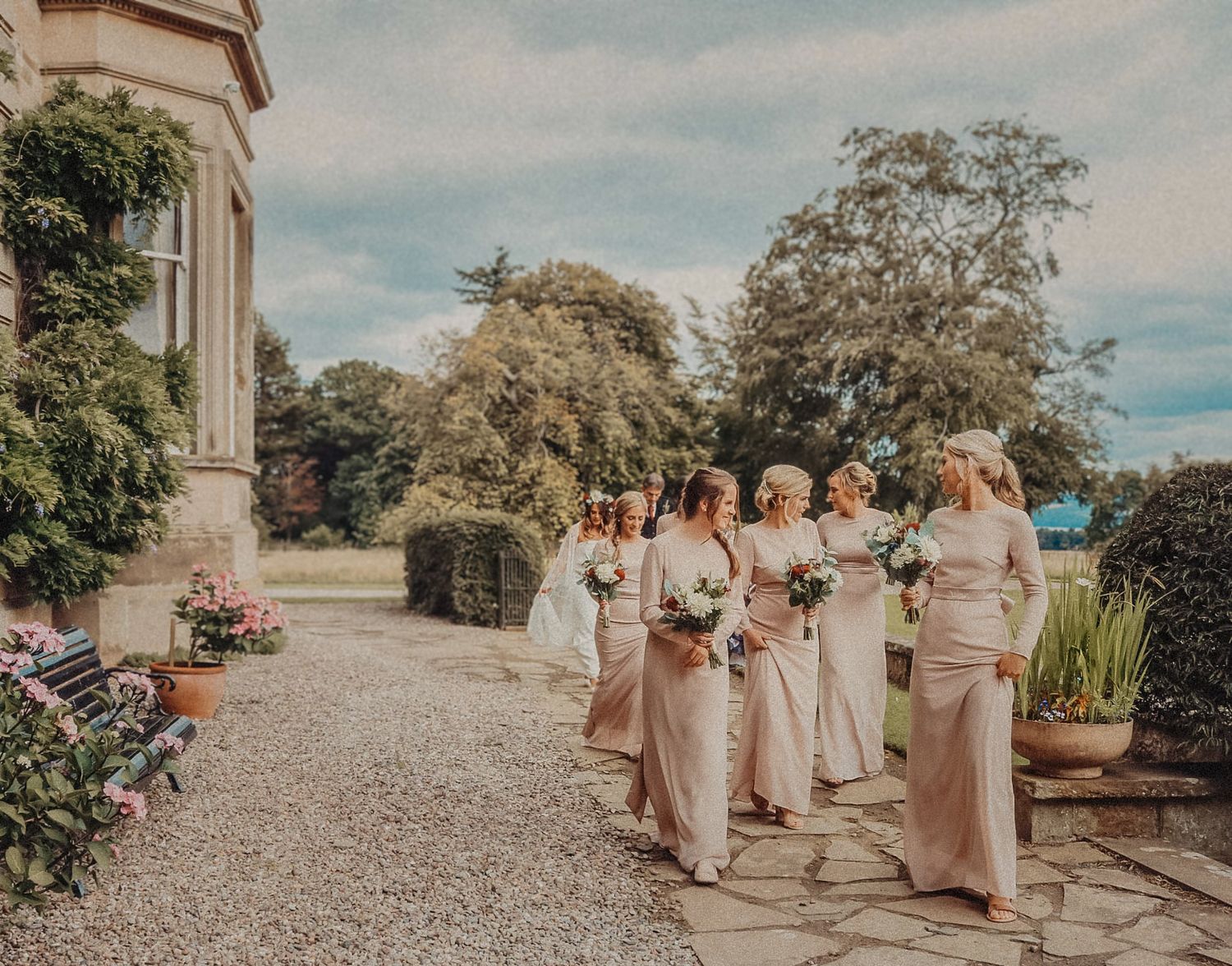 Wedding party in cream-colored dresses stands on a gravel path outside an elegant stone mansion with flowering gardens.
