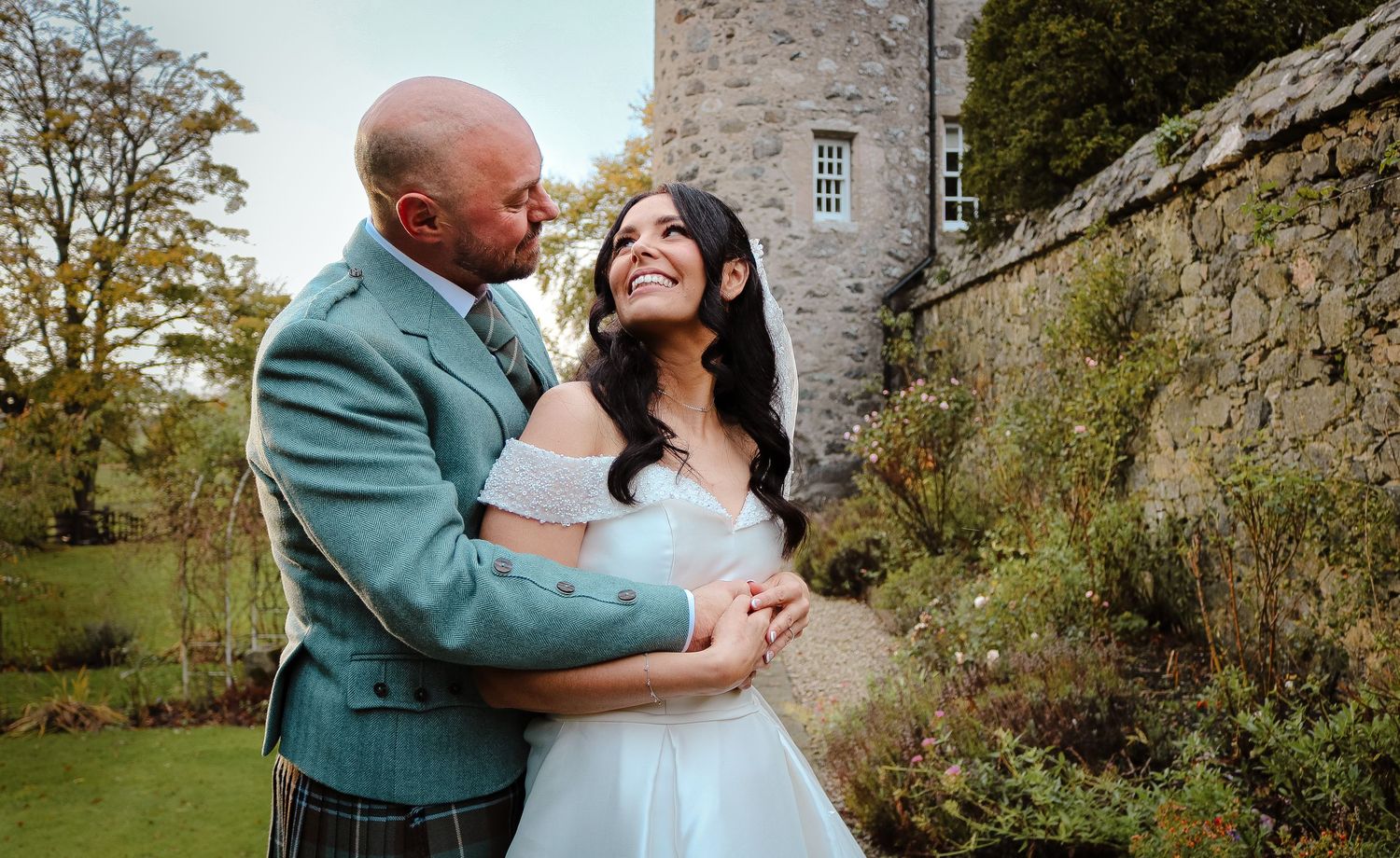 A joyful couple shares a romantic moment together in their wedding attire by a stone castle wall in a garden setting.