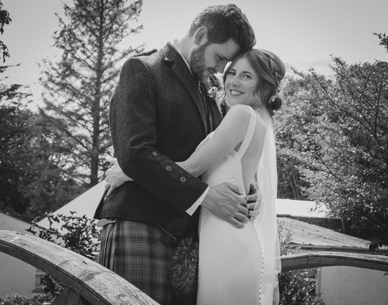 A romantic black and white wedding photo of a couple embracing on a bridge with evergreen trees in the background.
