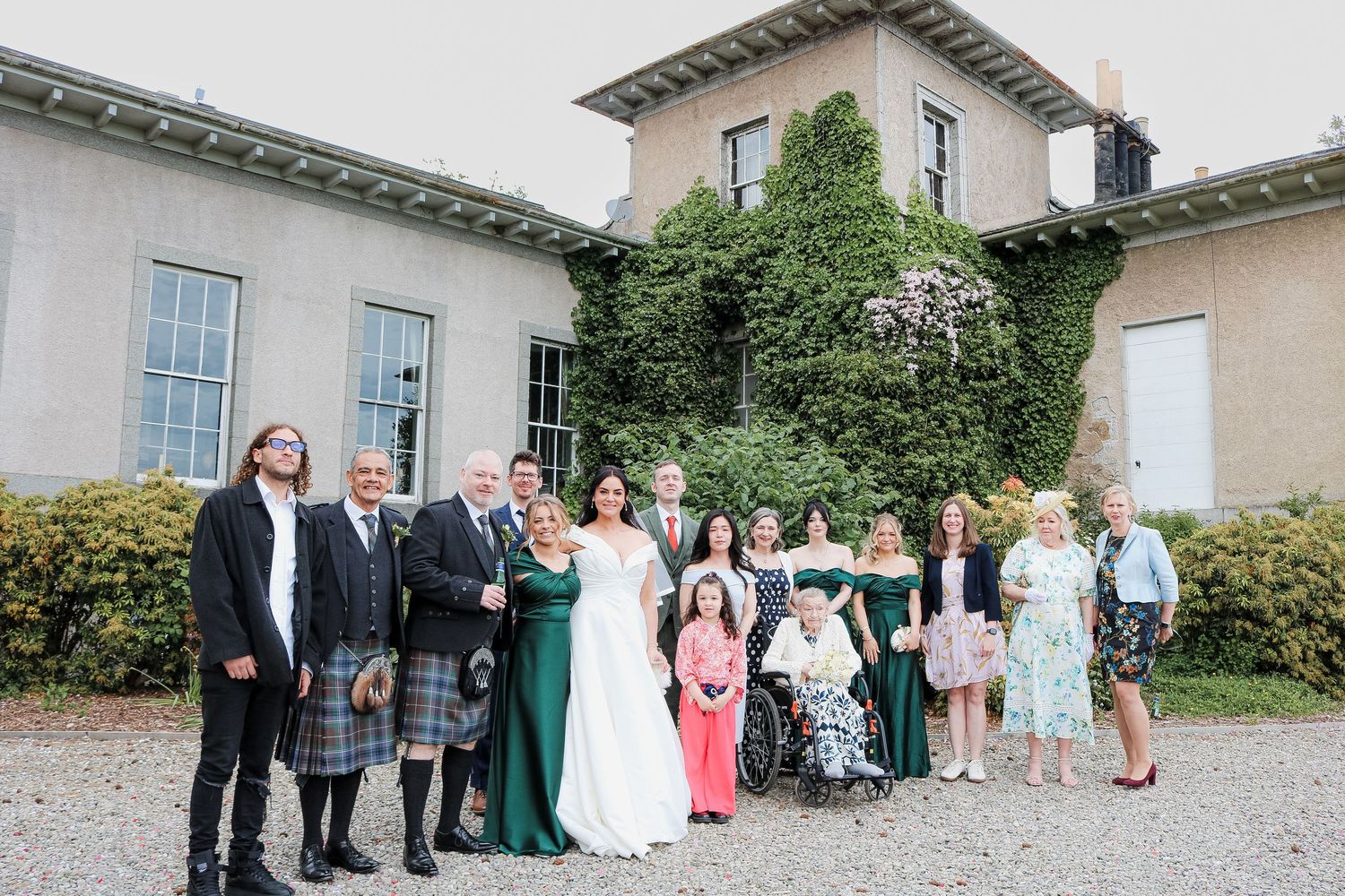 Wedding group photo outside an ivy-covered venue, bride in white gown with guests in formal attire including kilts.