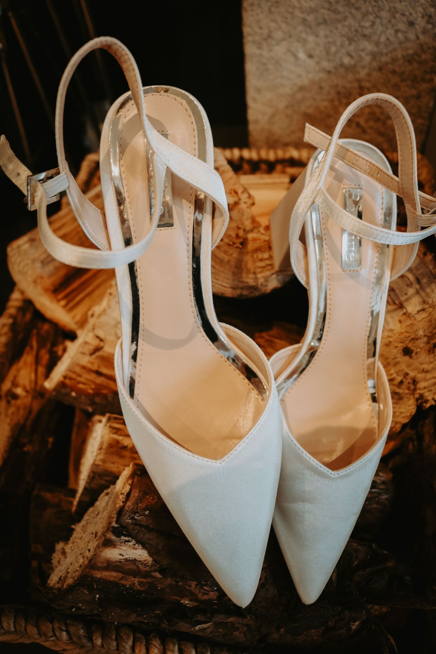 Elegant white pointed-toe bridal heels with delicate ankle straps displayed on rustic wooden surface.