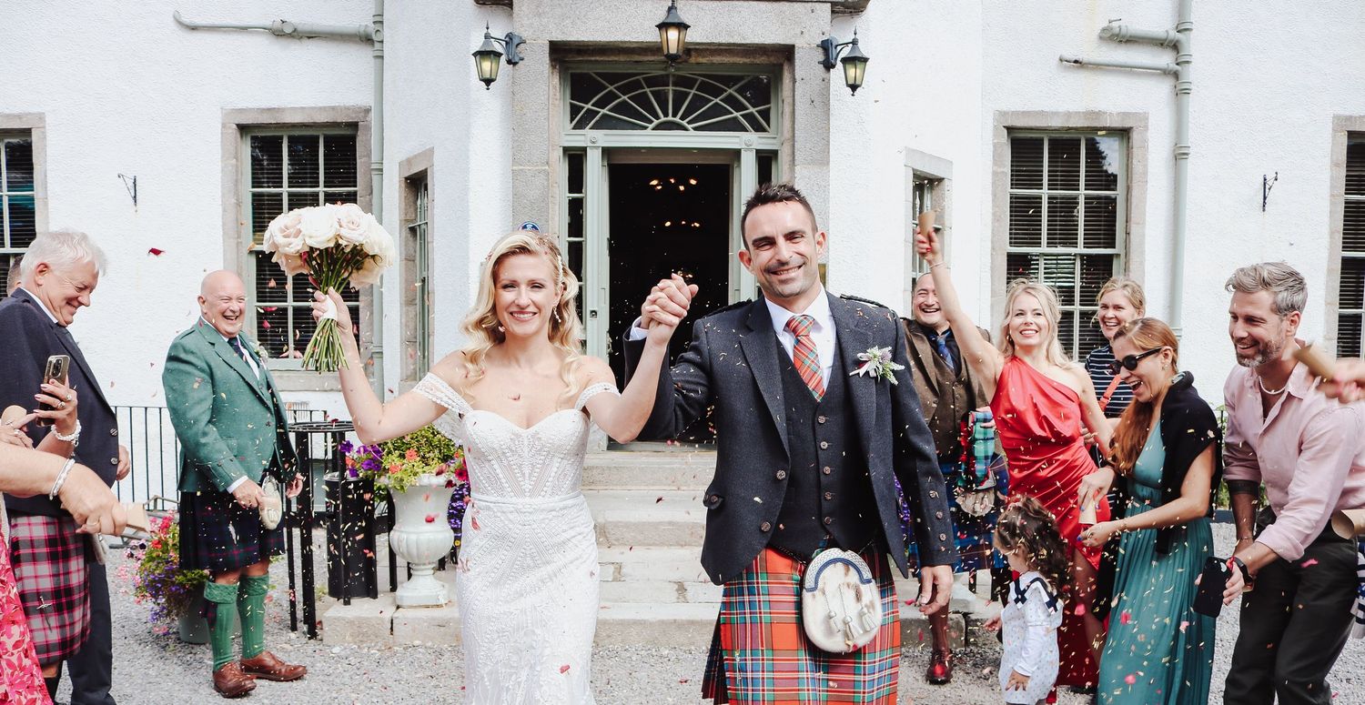 Wedding guests shower happy newlyweds with confetti outside white building as groom wears traditional Scottish kilt.