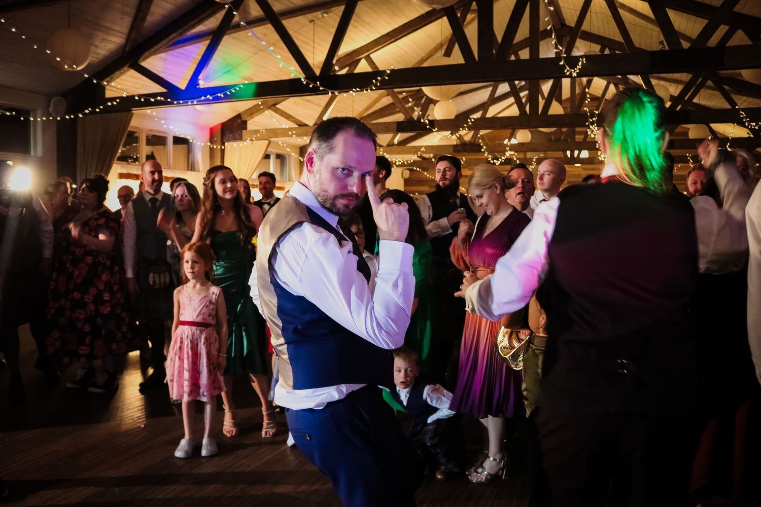 Wedding guests dancing and celebrating on a dimly lit dance floor illuminated by colorful party lights.