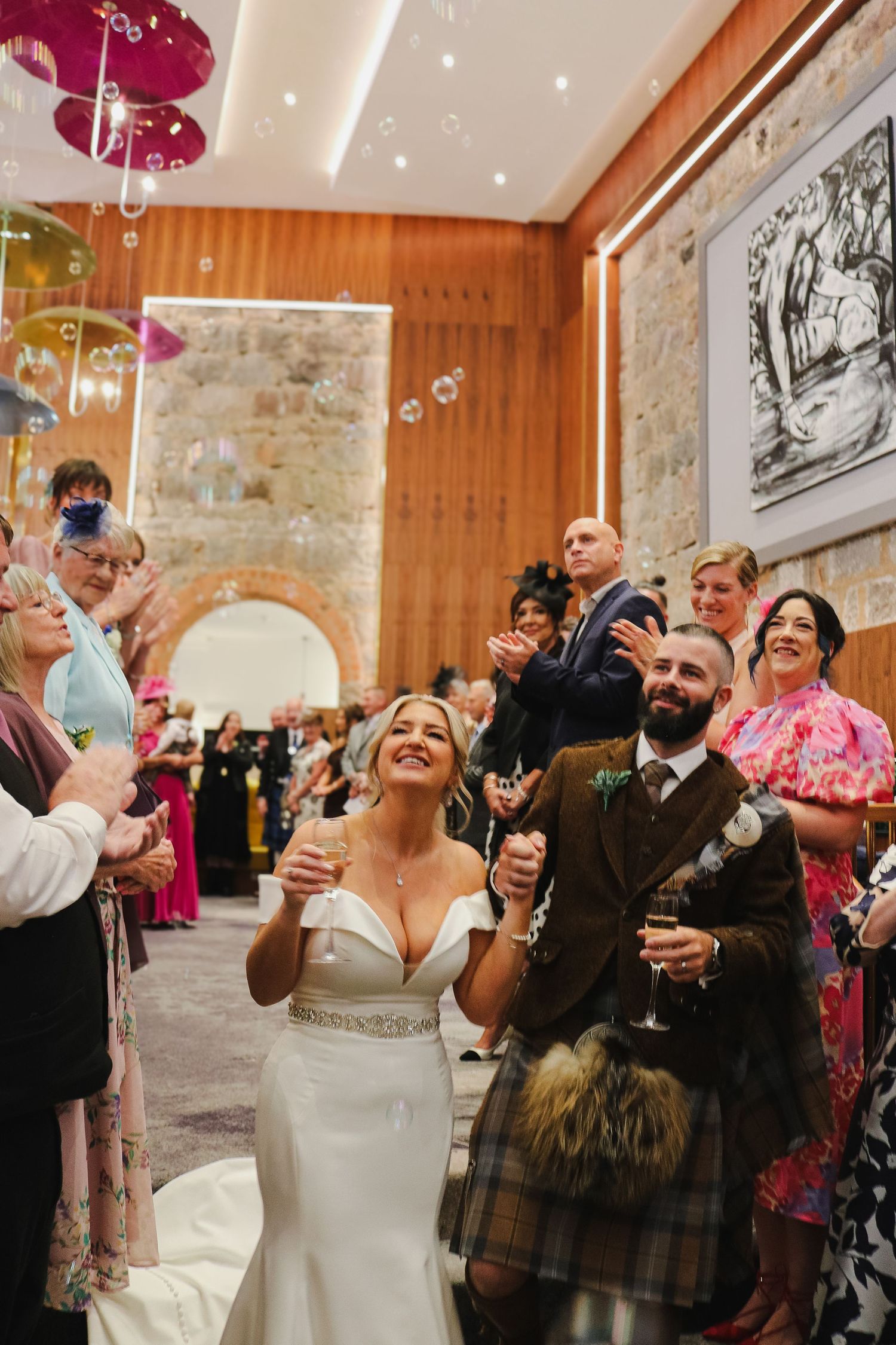 Wedding guests raise their glasses in celebration during a toast at an elegant reception with stone walls and wood paneling.