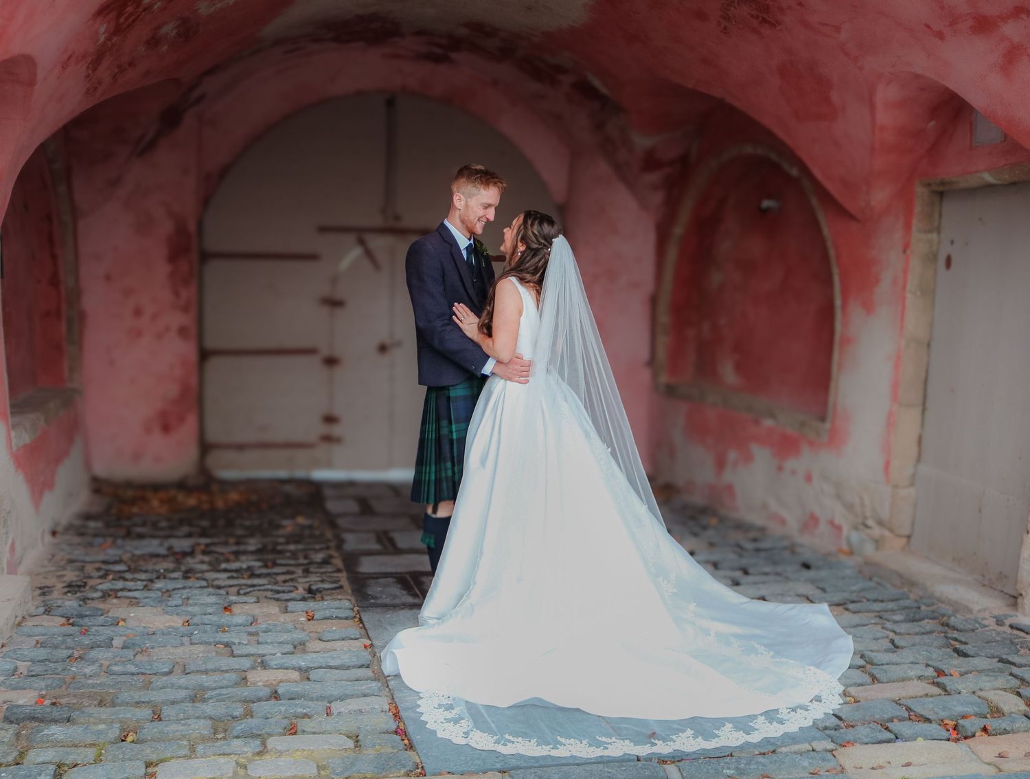 A bride in a flowing white gown and veil poses in a rustic stone corridor with peeling pink walls.