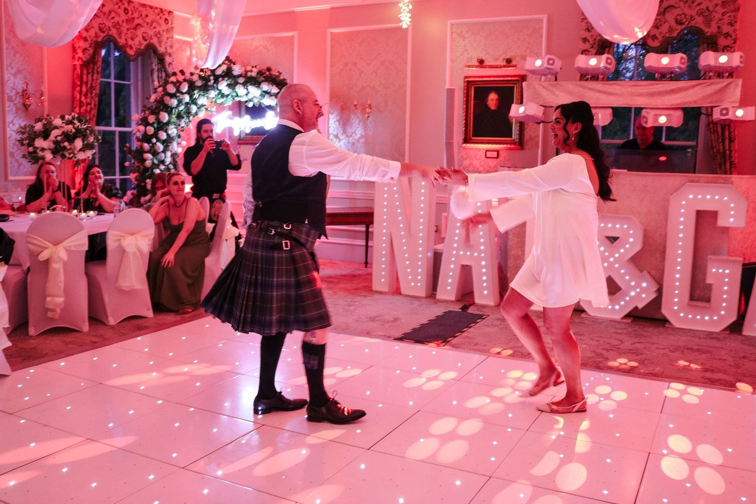 Couple dancing at a wedding reception on a white dance floor with illuminated letter decorations and pink lighting.