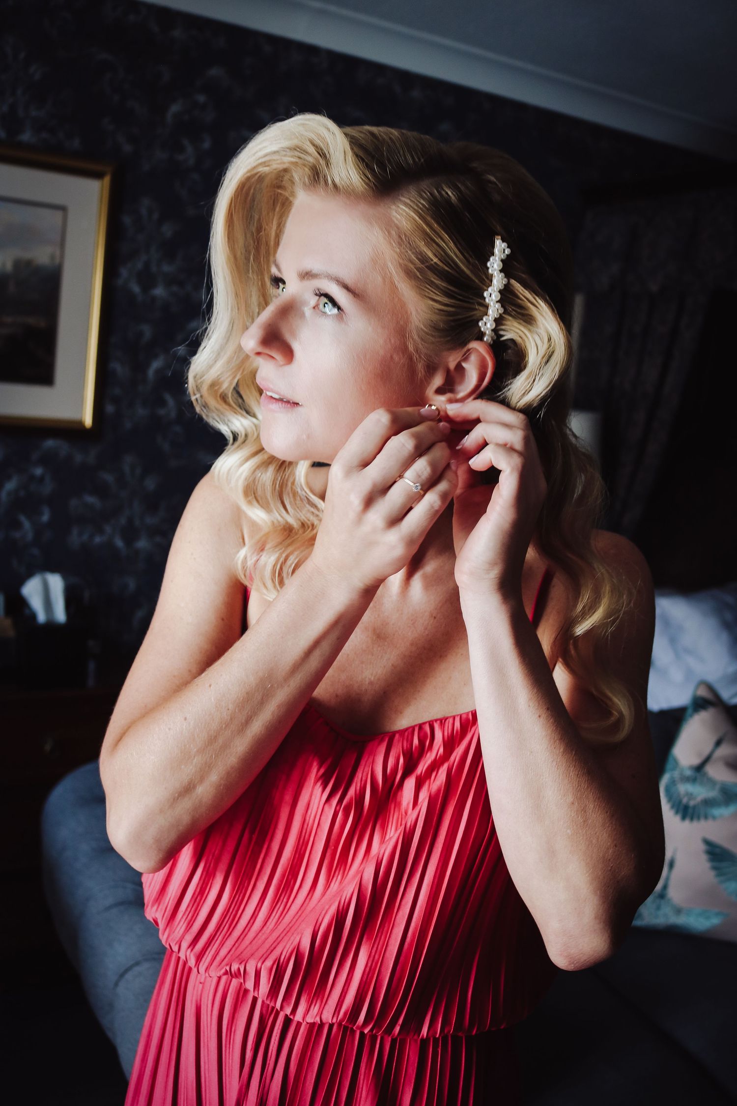 Someone in a coral pleated dress adjusts their earring while wearing vintage-inspired wavy blonde hair with sparkly hair clip.