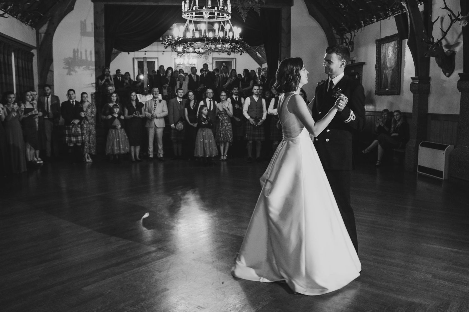 A couple shares their first dance as newlyweds under a crystal chandelier in an elegant ballroom with guests watching.