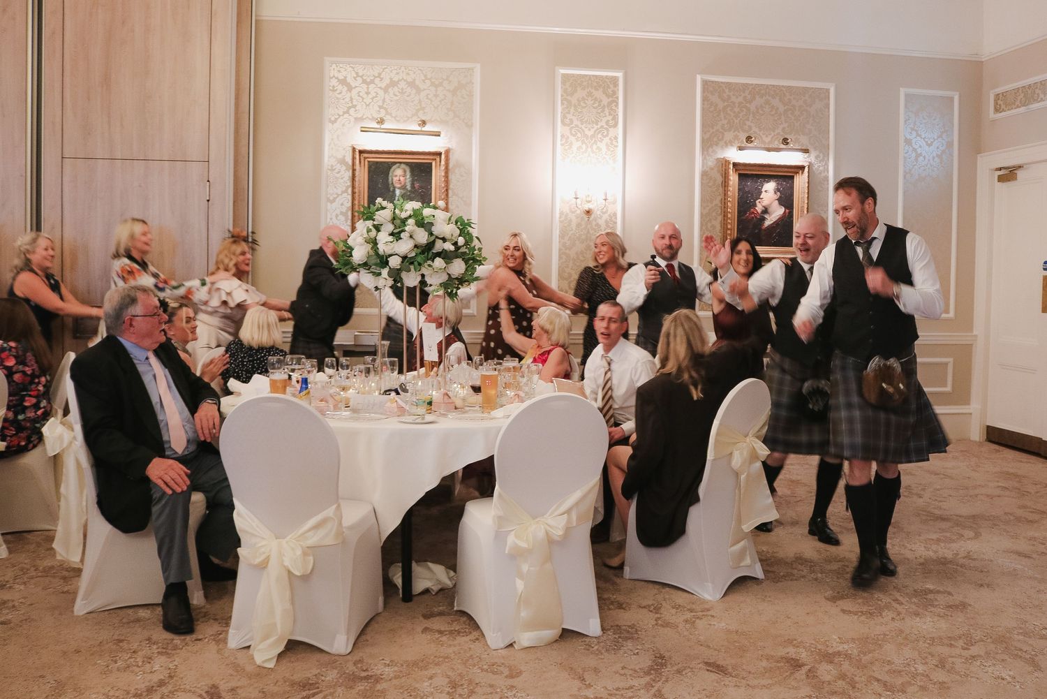 Wedding guests in formal attire seated at elegantly decorated tables in a grand reception hall, with a kilted groomsman.