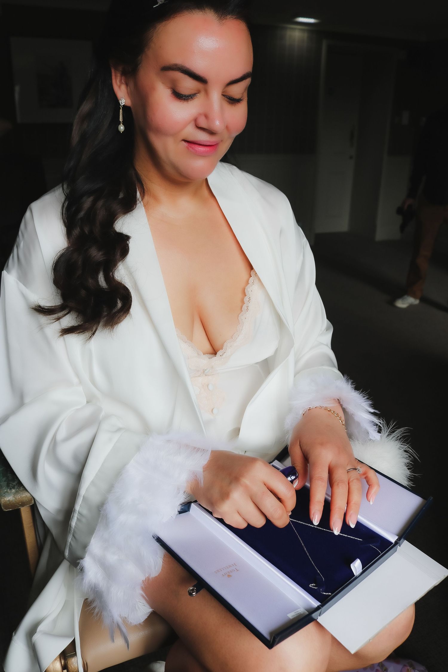 Bride in white robe opening a jewelry box, wearing pearl earrings and lace lingerie during wedding preparations.