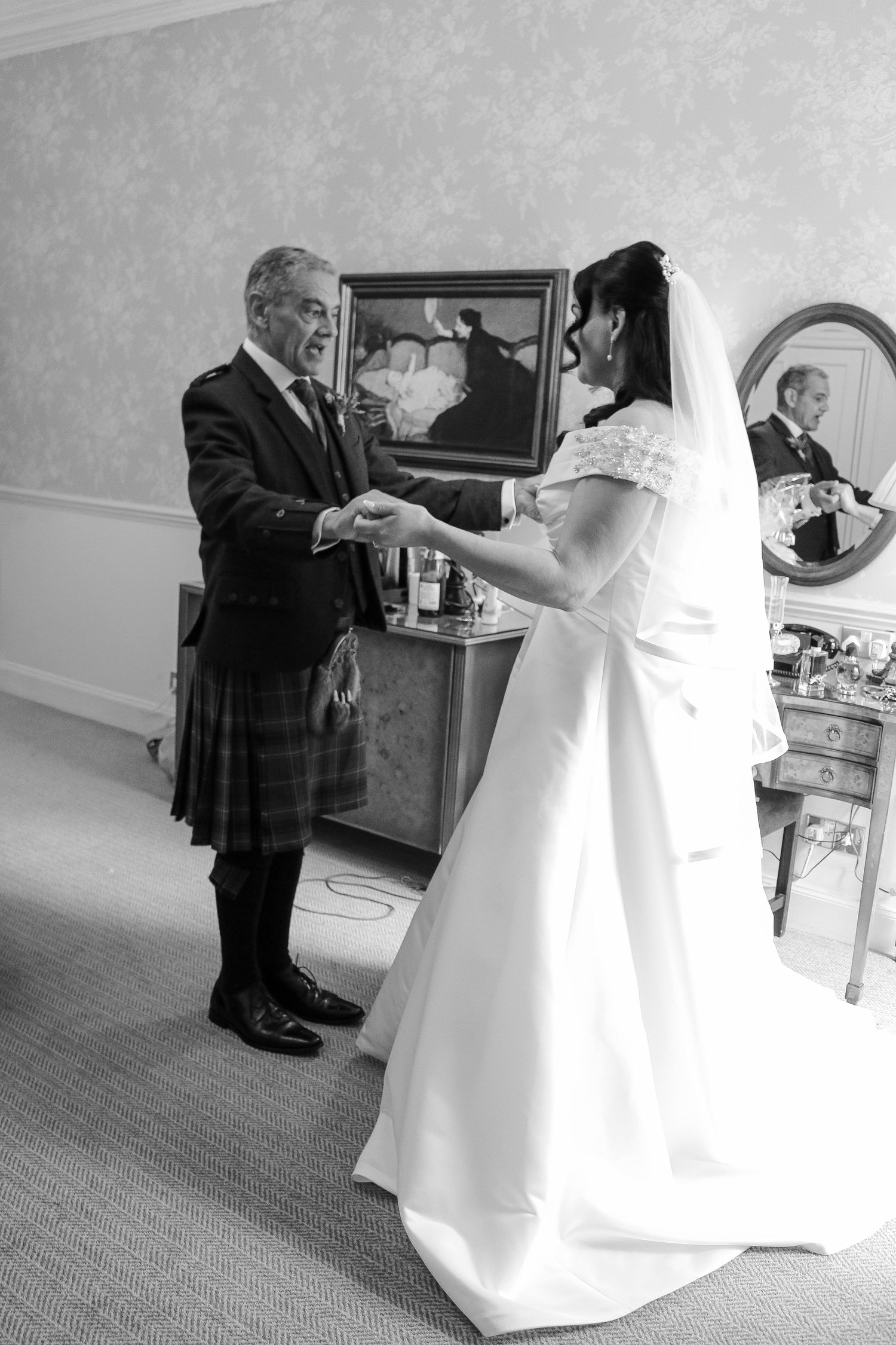 Black and white photo of a bride in white gown holding hands with a man in a kilt in an elegant room.