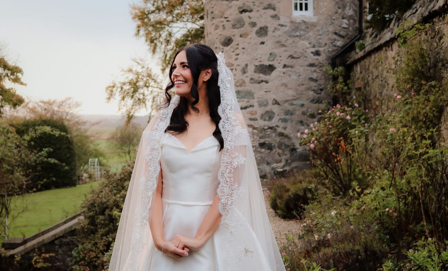 A flowing white wedding dress and veil against a rustic stone wall backdrop in an outdoor garden setting.