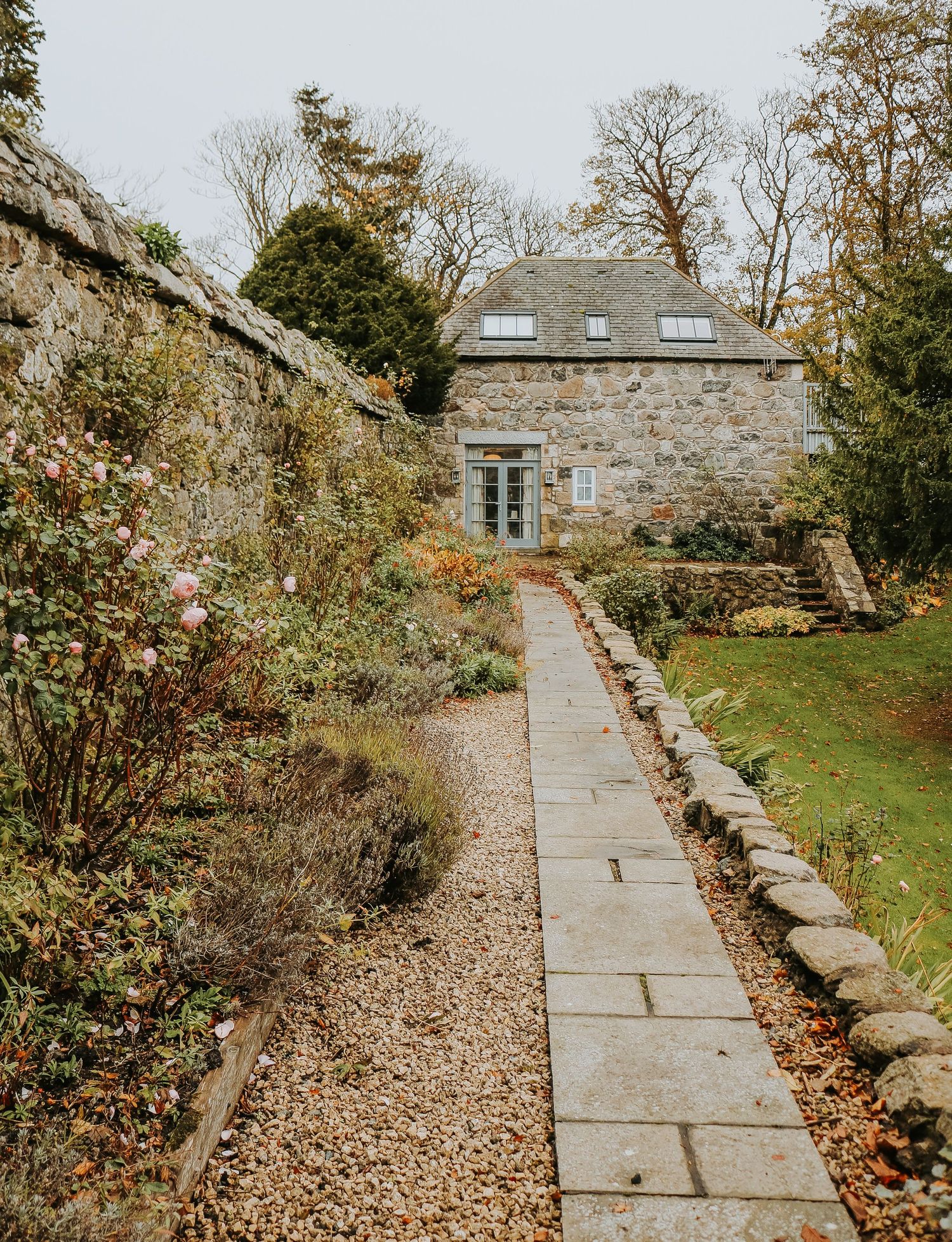 Stone walkway with autumn leaves leads to rustic stone buildings through a garden setting.