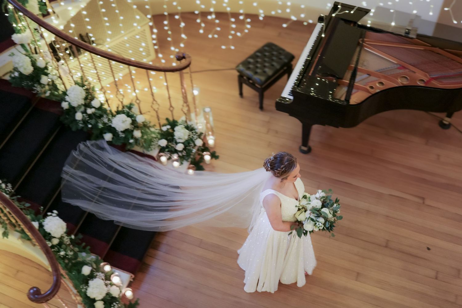 A flowing veil trails across a wooden floor decorated with white floral arrangements and a grand piano in an elegant setting.