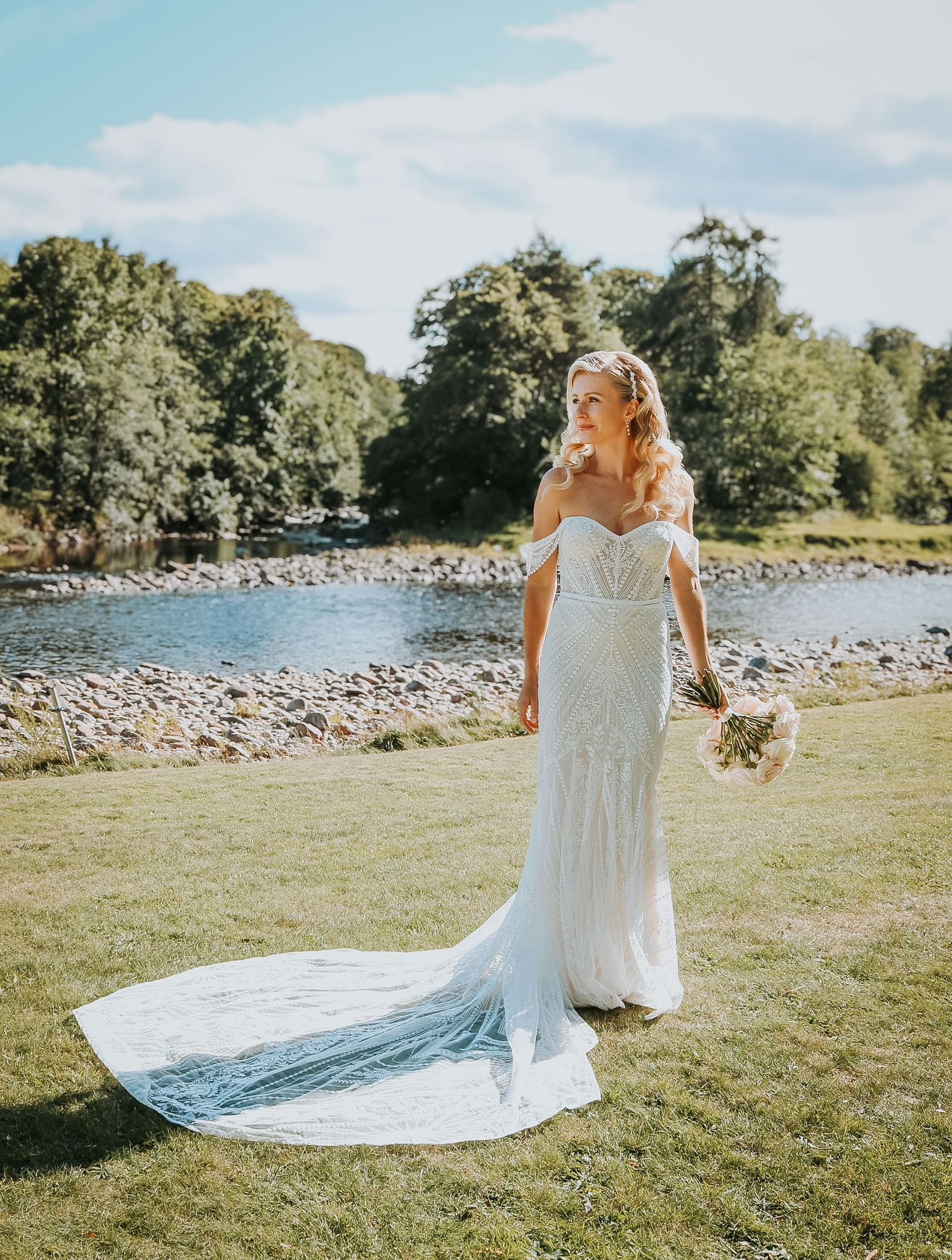 A bride in a flowing off-shoulder lace wedding gown with long train stands by a riverside on a sunny summer day.