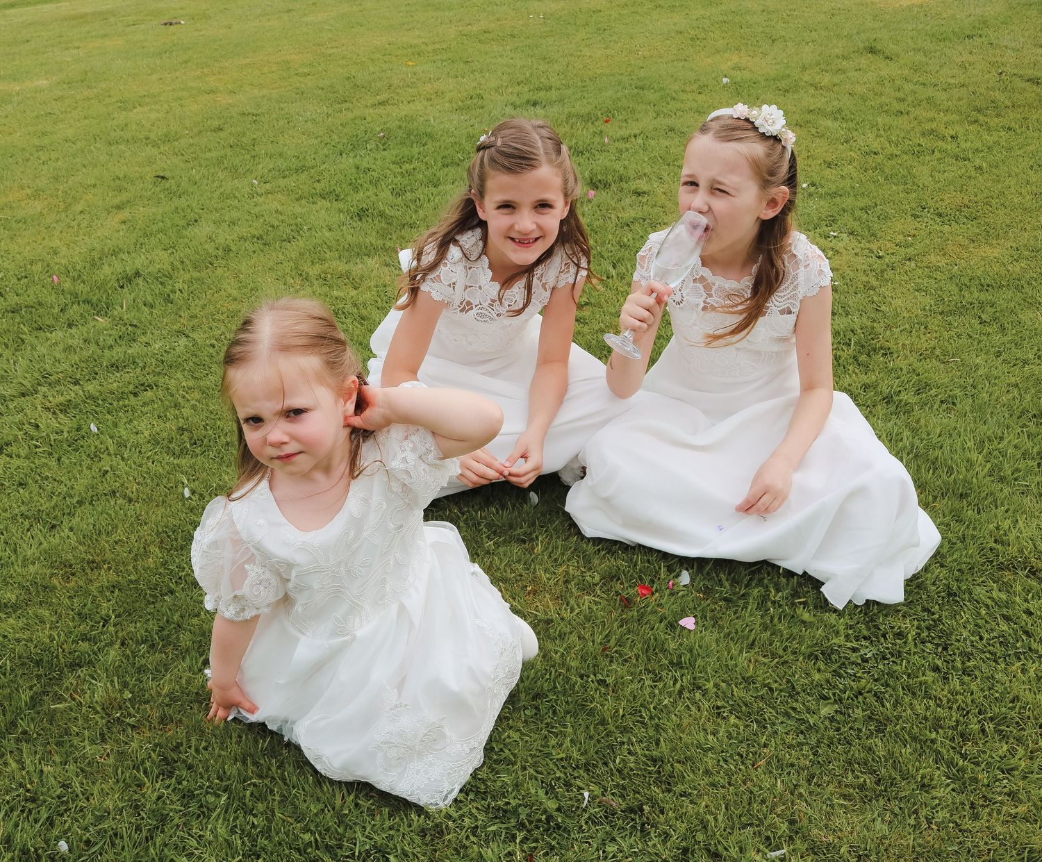 Three young girls in white dresses sitting on green grass, likely flower girls at a wedding celebration.