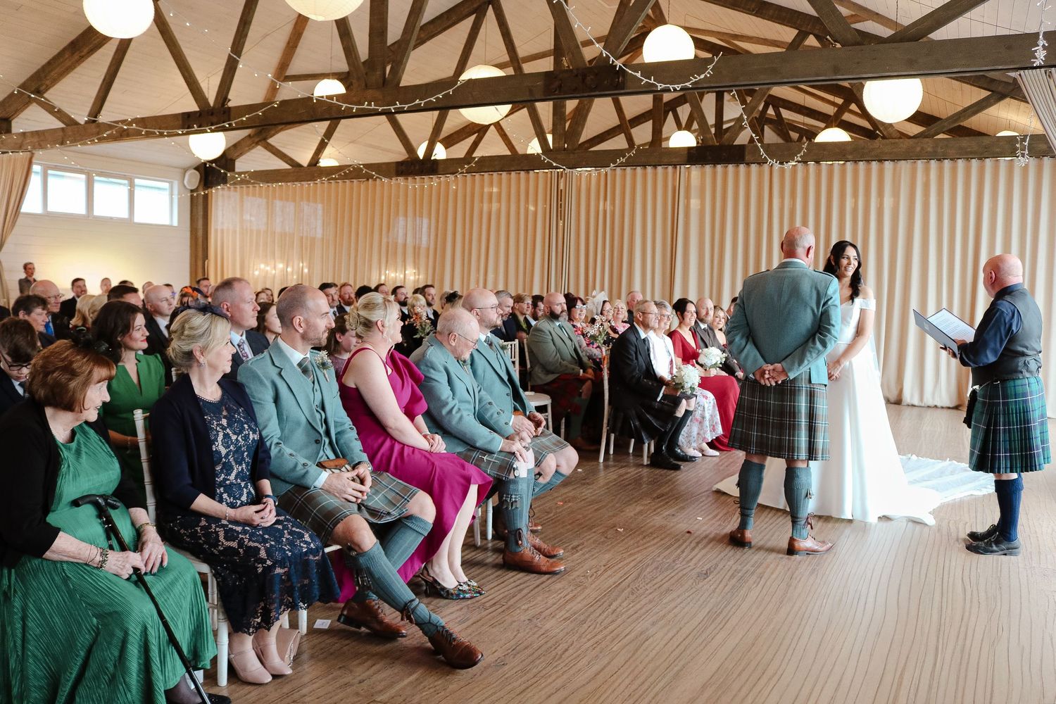 A traditional Scottish wedding ceremony takes place in a rustic wooden hall with guests seated in rows.