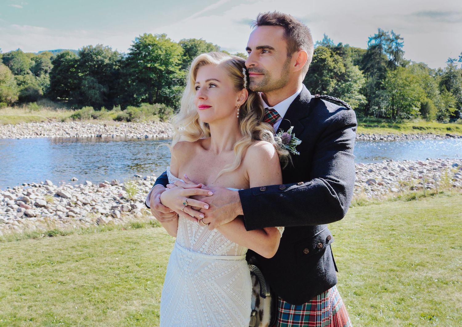 A romantic wedding couple embraces beside a lake on a sunny day, with the partner wearing a traditional kilt.