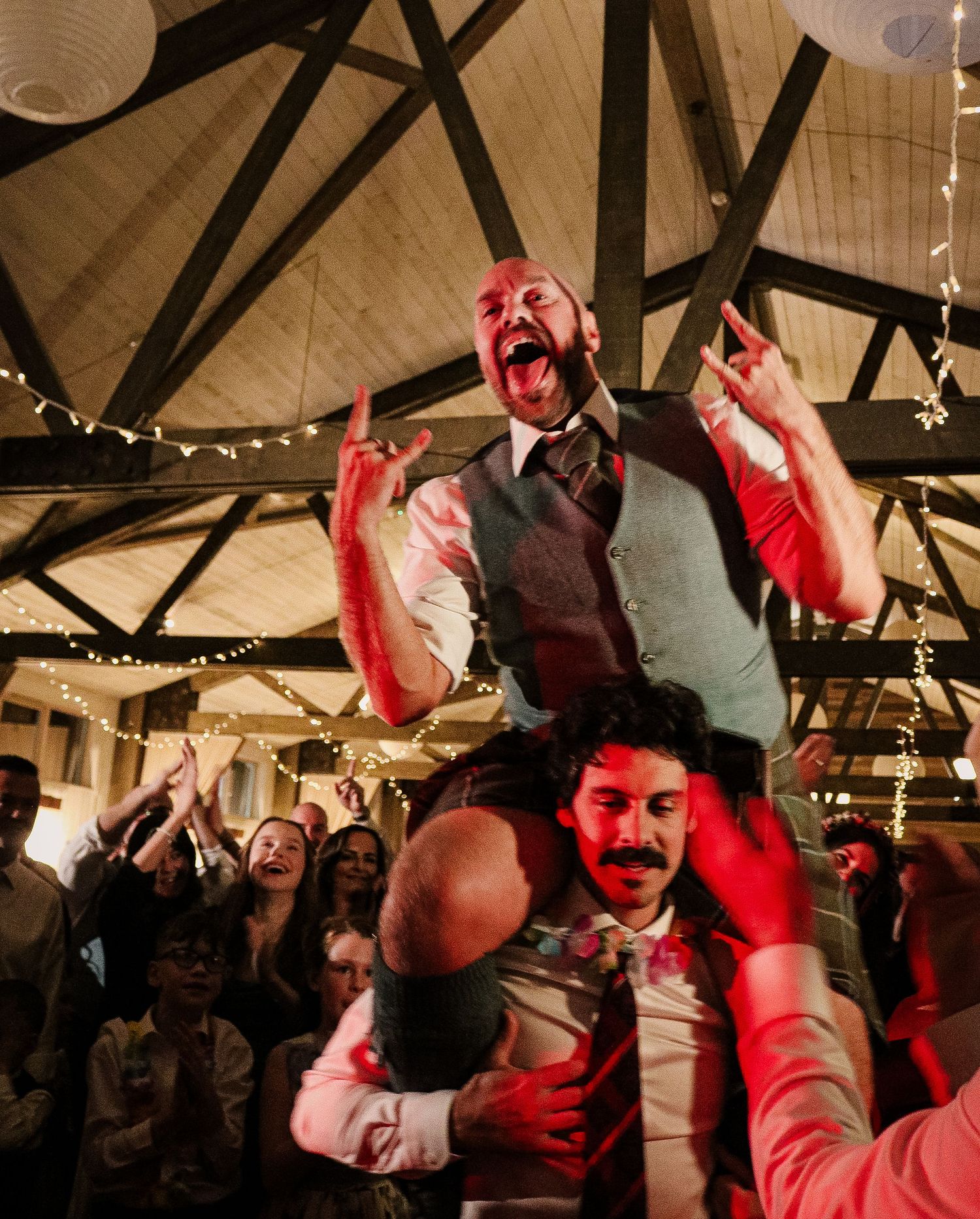 People dancing and celebrating at a festive indoor venue with string lights and exposed wooden beams overhead.
