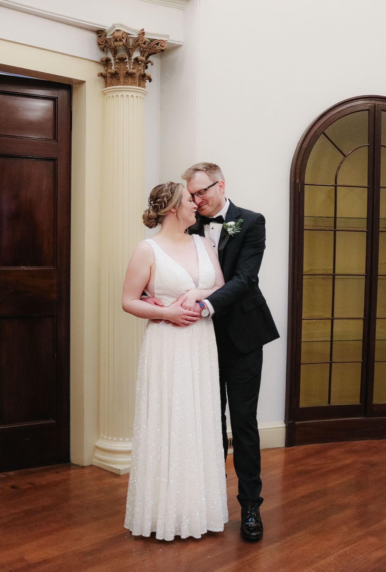 Elegant couple in wedding attire share an intimate moment near ornate column and arched doorway in classic interior.