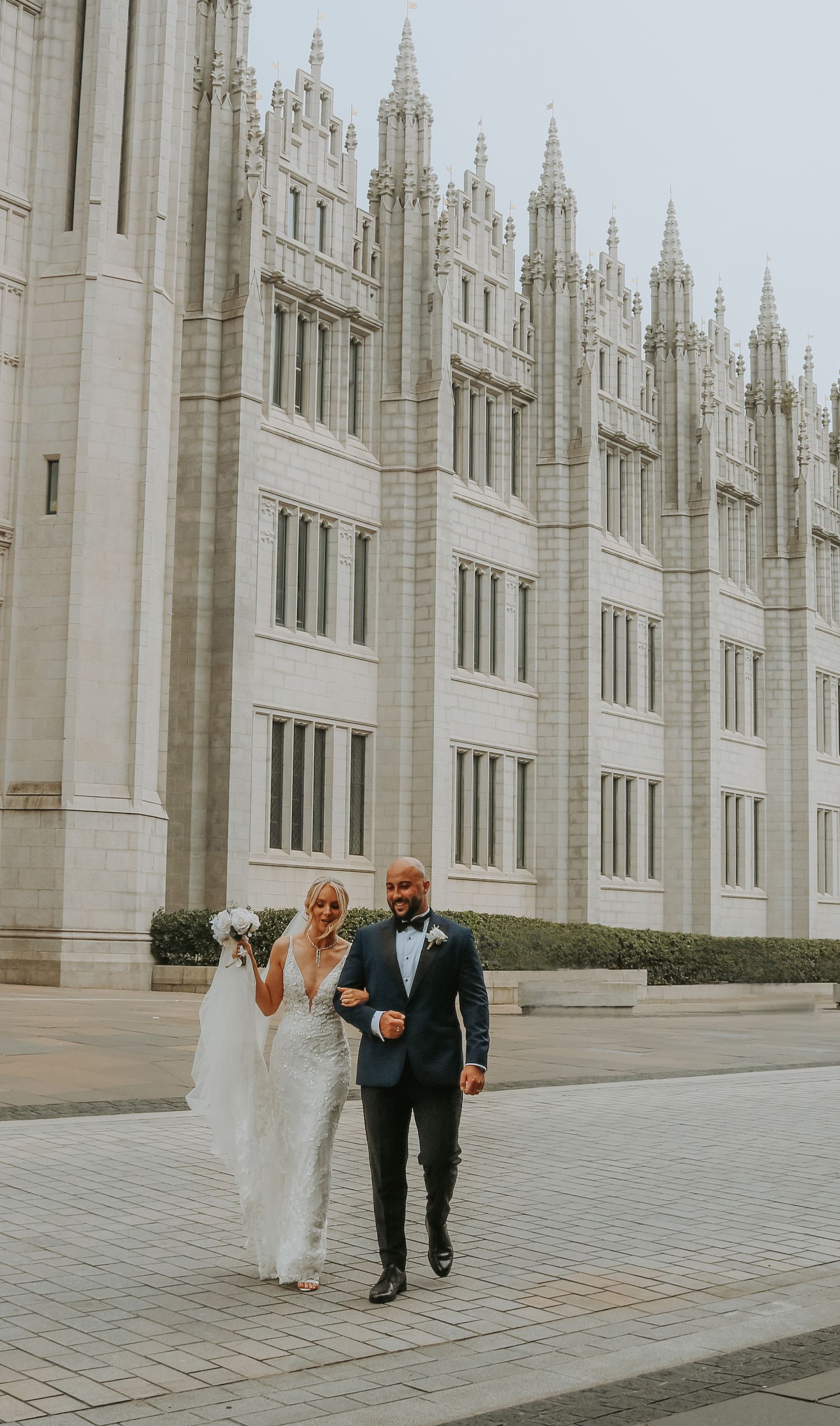 A couple walks arm in arm outside the ornate Gothic architecture of the Salt Lake Temple on their wedding day.