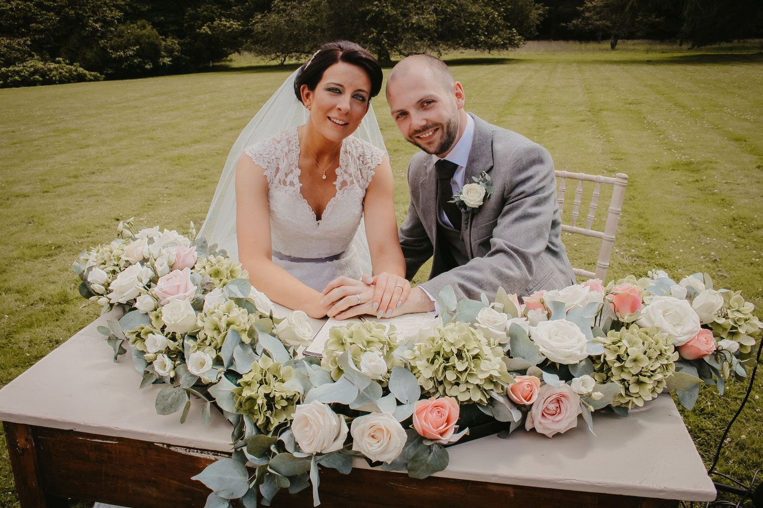 A wedding couple sits at a rustic wooden table adorned with a lush floral arrangement of pink roses and eucalyptus.